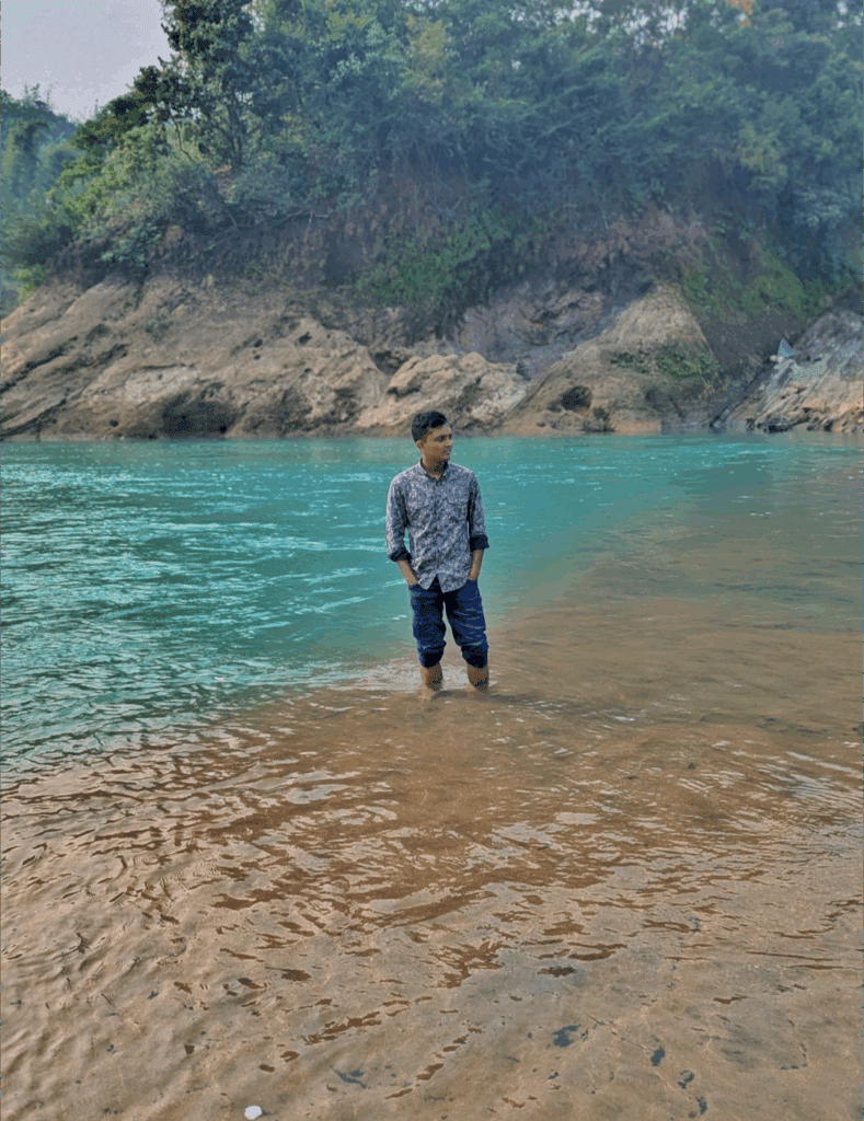 A male traveler standing in the emerald waters of the Lalakhal River in Sylhet with hilly terrain in the background.